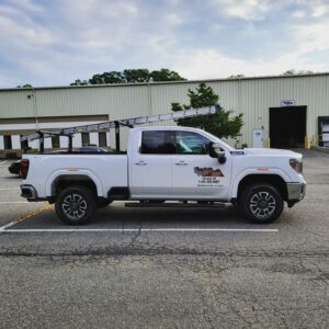 A white pickup truck with a ladder on top is parked in a lot outside a beige industrial building. The truck's door has a decal with the Tech Roofing logo and phone number of a roof service company.